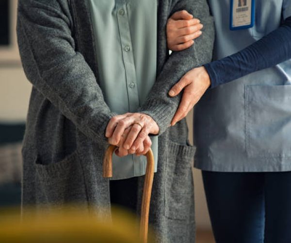 Close up hands of caregiver doctor helping old woman at private clinic. Close up of hands of nurse holding a senior patient with walking stick. Elder woman using walking cane at nursing home with nurse holding hand for support.